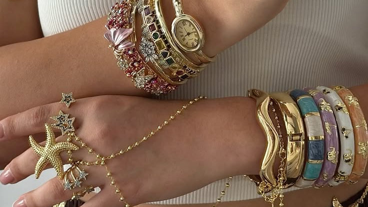 A person wearing a coordinated gold jewelry set featuring starfish and shell motifs, including a waist chain, bracelets, rings, and layered necklaces, paired with a white ribbed tank top and brown pleated skirt.